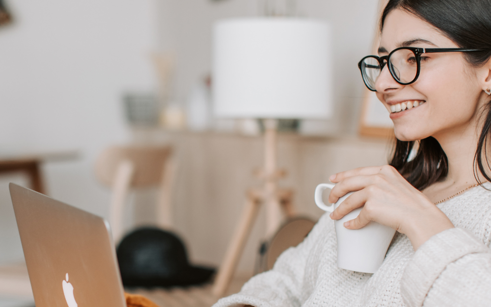 Persoon met bril werkt aan een laptop met een tas koffie in de hand.